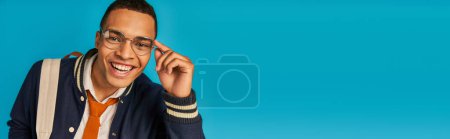 african american student in eyeglasses and jacket smiling at camera on blue, banner, copy space