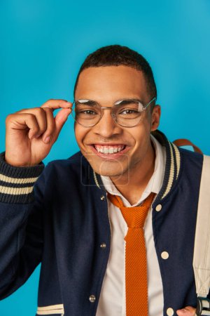 cheerful and trendy african american student in jacket, tie and eyeglasses smiling at camera on blue