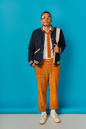 confident african american student in jacket and orange pants holding hand in pocket on blue
