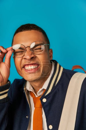 funny african american student in jacket adjusting eyeglasses and grimacing on blue background