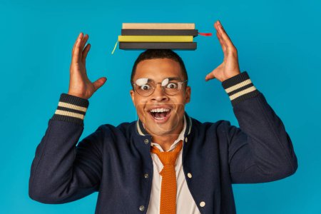 amazed african american student posing with notebooks on head on blue, eyeglasses, stylish jacket