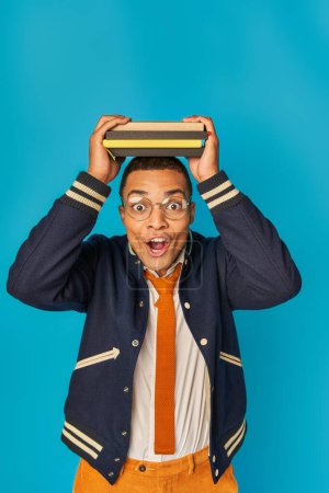 astonished african american student in trendy jacket and eyeglasses with notebooks on head on blue