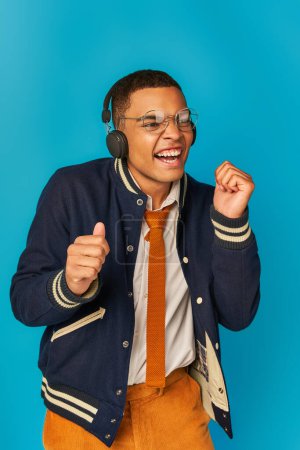 estudiante afroamericano lleno de alegría en chaqueta de moda escuchando música en auriculares inalámbricos en azul