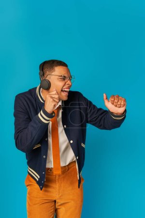 Foto de Energizado, elegante estudiante afroamericano en auriculares inalámbricos escuchando música en azul - Imagen libre de derechos