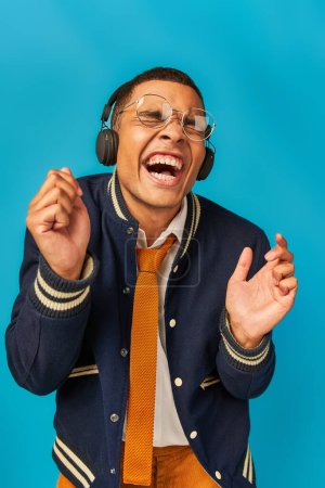überglücklicher, trendiger afrikanisch-amerikanischer Student mit drahtlosen Kopfhörern, der mit geschlossenen Augen auf blau lacht