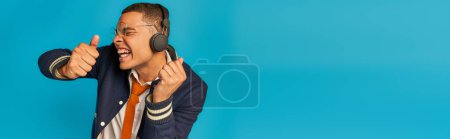 Téléchargez les photos : Étudiant afro-américain excité dans les écouteurs sans fil montrant pouces vers le haut sur bleu, bannière - en image libre de droit