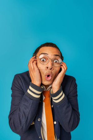 Schockierter afrikanisch-amerikanischer Student mit Brille und Kopfhörer blickt auf blauem, offenem Mund in die Kamera