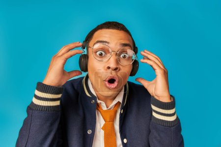 estudiante afroamericano impresionado en gafas y auriculares, con la boca abierta en azul