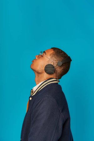Téléchargez les photos : Vue latérale d'un étudiant afro-américain branché en veste écoutant de la musique dans un casque sur bleu - en image libre de droit