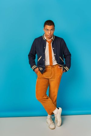Téléchargez les photos : Étudiant afro-américain confiant en veste et pantalon orange posant avec les mains dans les poches sur bleu - en image libre de droit