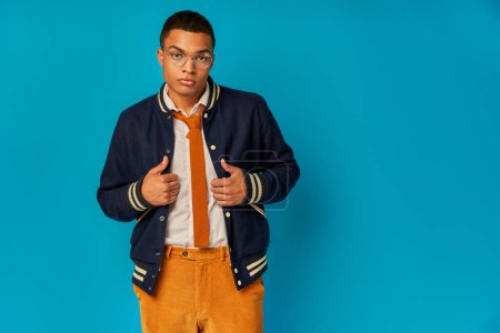 confident african american student in trendy jacket and orange pants looking at camera on blue