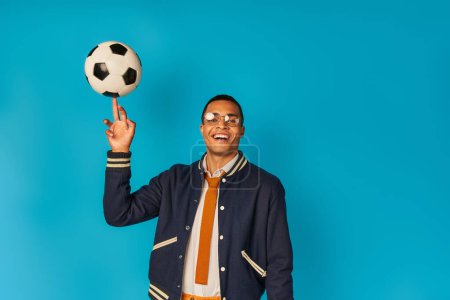 fröhliche und stylische afrikanisch-amerikanische Studentin spielt mit Fußball und schaut in die Kamera auf blau