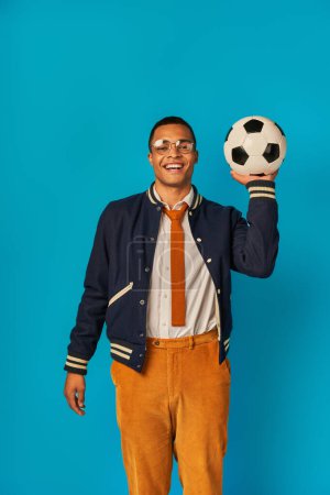 estudiante afroamericano feliz en chaqueta y pantalones anaranjados jugando con pelota de fútbol en azul