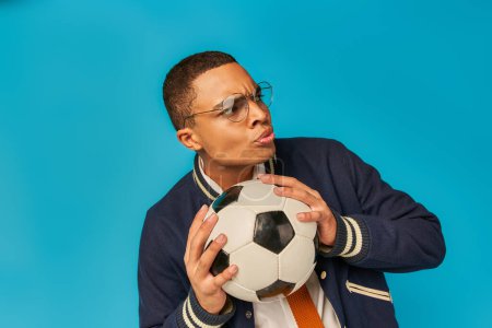 serio africano americano estudiante en gafas celebración fútbol pelota y mirando hacia otro lado en azul