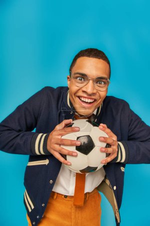 überglücklicher Afroamerikaner in Jacke und Brille, Fußballball in der Hand, lächelnd in die Kamera auf blau