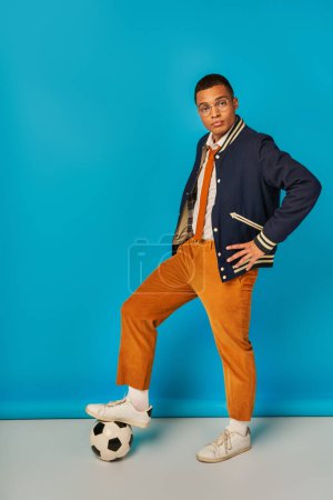 estudiante afroamericano de moda en chaqueta, pantalones anaranjados pisando pelota de fútbol en azul, mano en la cadera