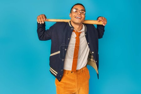 Photo for Happy and stylish african american student with baseball bat looking at camera on blue background - Royalty Free Image