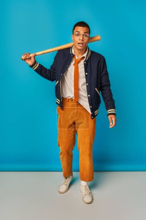 expressive african american student in orange pants holding baseball bat, looking at camera on blue