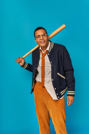 african american student in orange pants holding baseball bat and smiling at camera on blue