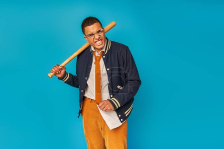 wütender afrikanisch-amerikanischer Student in stylischer Kleidung mit Baseballschläger und Fratze auf blauem Grund