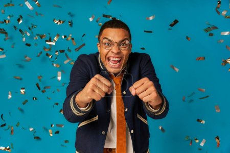 Téléchargez les photos : Ravi et tendance afro-américain étudiant riant lors de la fête sous confettis tombant sur bleu - en image libre de droit