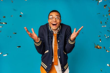 étudiant afro-américain joyeux et à la mode montrant wow geste lors de la fête sous confettis sur bleu