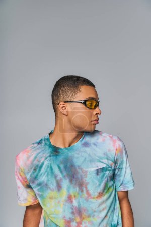youthful african american man in sunglasses and tie-dye t-shirt looking away on grey