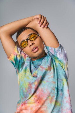 african american man with hands above head looking at camera on grey, sunglasses, tie-dye t-shirt
