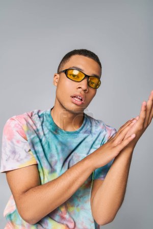 joven, hombre afroamericano con estilo en camiseta tie-dye y gafas de sol mirando a la cámara en gris