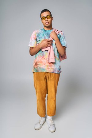 trendy african american man, sunglasses, tie-dye t-shirt, orange pants on grey, full length