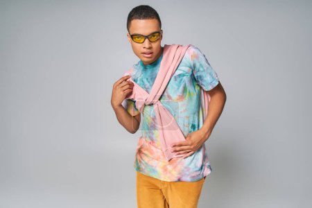 Photo for African american man in sunglasses and tie-dye t-shirt posing with hand on hip on grey - Royalty Free Image