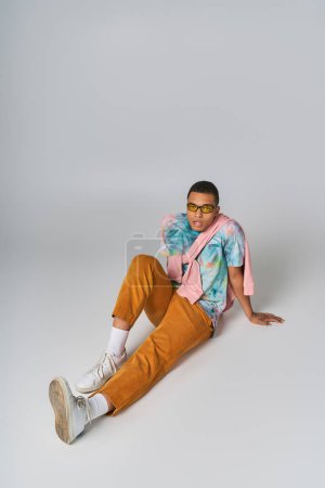 trendy african american man sitting on grey, sunglasses, orange pants, tie-dye t-shirt