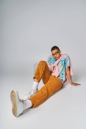 Foto de Hombre afroamericano en camiseta de tie-dye, pantalones naranjas y gafas de sol sentados en gris - Imagen libre de derechos