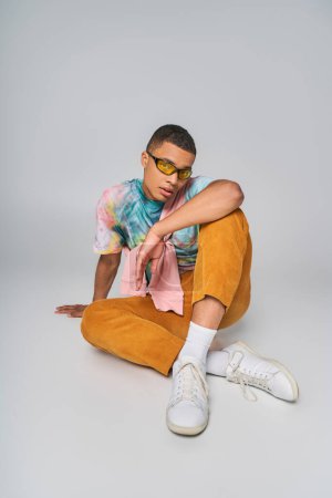 hombre afroamericano moderno en gafas de sol, pantalones anaranjados, corbata-tinte camiseta sentado en gris