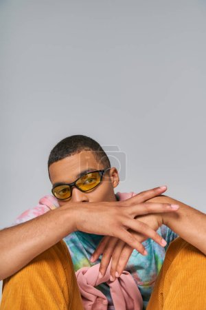 Téléchargez les photos : Jeune homme afro-américain en lunettes de soleil à la mode et t-shirt tie-dye regardant la caméra sur gris - en image libre de droit