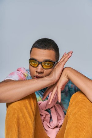 trendy african american guy in trendy sunglasses and tie-dye t-shirt looking at camera on grey