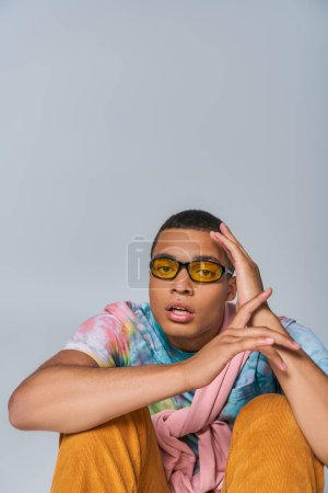 youthful african american man in trendy sunglasses and tie-dye t-shirt looking at camera on grey