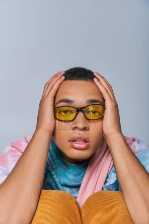 Foto de Hombre afroamericano de moda en gafas de sol, tie-dye camiseta tocando la cabeza, mirando a la cámara en gris - Imagen libre de derechos