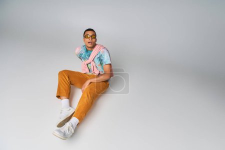 Téléchargez les photos : Homme afro-américain en pantalon orange, t-shirt tie-dye, lunettes de soleil, regardant caméra sur gris - en image libre de droit