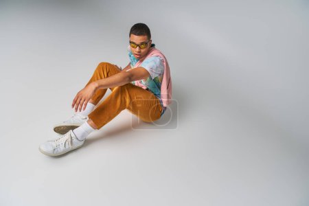 Foto de Hombre afroamericano, pantalones anaranjados, camiseta tie-dye, gafas de sol sentadas en gris, mirando a la cámara - Imagen libre de derechos