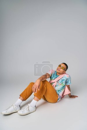 chico afroamericano, pantalones anaranjados, corbata-tinte camiseta, gafas de sol sentado en gris, mirando hacia otro lado