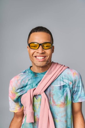 happy african american man in sunglasses and tie-dye t-shirt looking at camera on grey