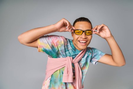 fröhlicher afrikanisch-amerikanischer Mann in Sonnenbrille und Krawatten-T-Shirt lächelt in die Kamera auf grau