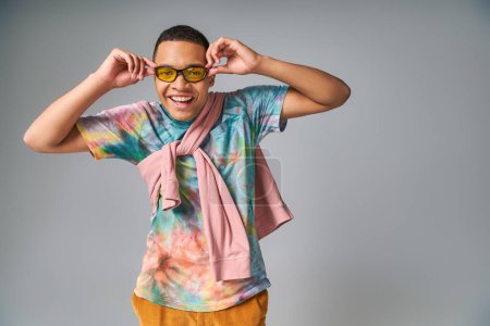 happy african american man in tie-dye t-shirt adjusting sunglasses and smiling at camera on grey