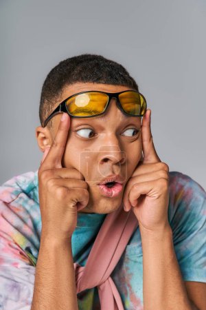 Téléchargez les photos : Surpris homme afro-américain avec des lunettes de soleil élégantes regardant loin sur gris - en image libre de droit