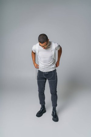 Modetrend, afrikanisch-amerikanischer Mann mit Sonnenbrille, T-Shirt, Jeans mit Händen in Taschen auf grau