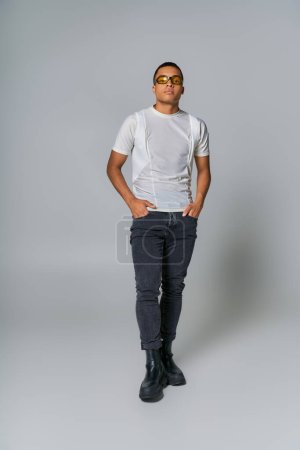 trendy african american man in sunglasses, t-shirt, jeans looking at camera on grey