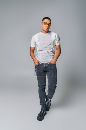 stylish african american man in sunglasses, t-shirt, jeans looking at camera on grey