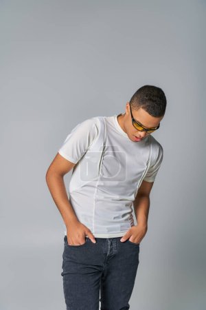 Foto de Joven hombre afroamericano en gafas de sol de moda, camiseta, jeans, con las manos en los bolsillos en gris - Imagen libre de derechos