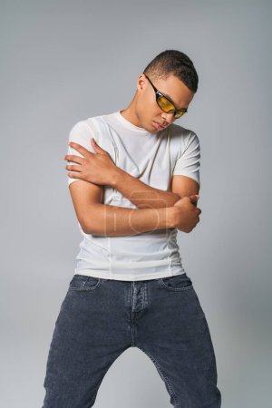joven hombre afroamericano en elegante camiseta, jeans y gafas de sol con los brazos cruzados en gris